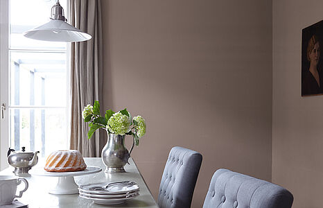 1759794_main_image_6922928fdd4c9 Nobles Erdbraun. Eine in diesem ruhigen und warmen Braun gestrichene Wand, davor ein Esstisch mit graublauen Polsterstühlen.