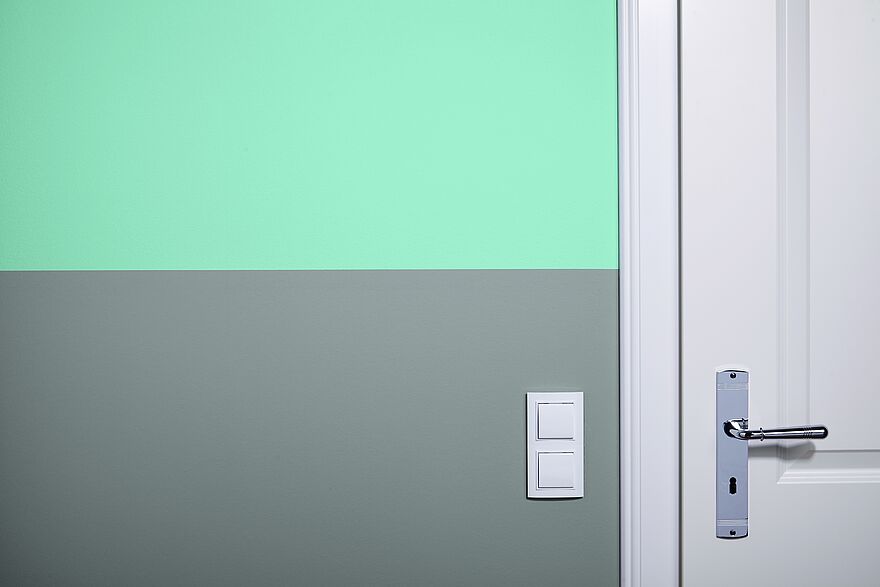 alpina-artikel-waende-zweifarbig-streichen-farbkante-header Eine zweifarbig gestrichene Wand mit einem grünen Farbstreifen oben und einem grauen Farbstreifen unten.