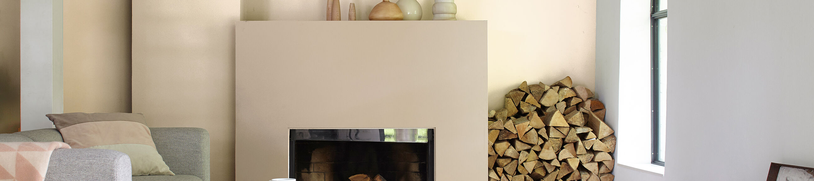 Graues Sofa vor beiger Wand und beigem Kamin, daneben ein Stapel Holzscheite